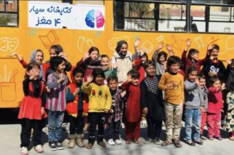 A group of Afghan children stand in front of a yellow bus, smiling. The bus has colorful drawings and text on it.