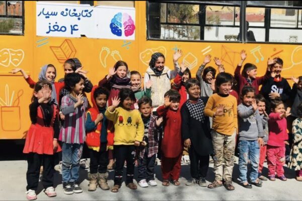 A group of Afghan children stand in front of a yellow bus, smiling. The bus has colorful drawings and text on it.