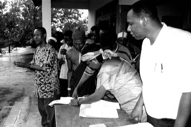 A woman leans over a table to sign a legal document, surrounded by community members looking on.