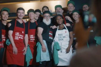 A group of chefs from June's HIV+ Eatery stand smiling together with red and white aprons.