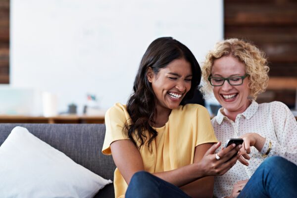 Two women on a couch laughing at something on a phone.