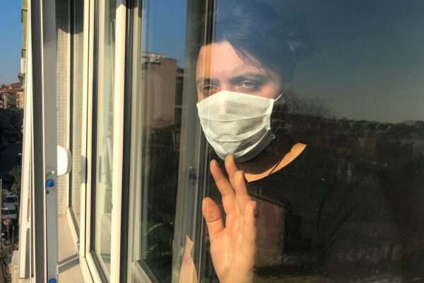 A young man wearing a mask stands looking out the window