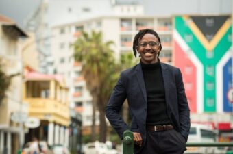 A South African man stands in front of buildings and the flag