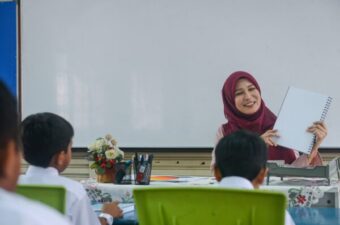 Asian teacher wearing a head covering, sitting at her desk smiling at children in the classroom.