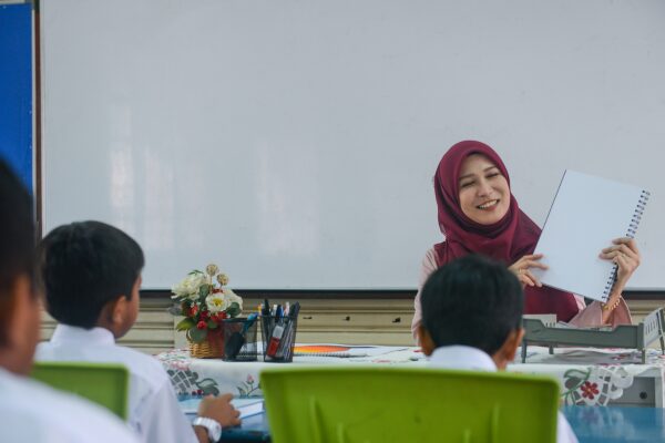 Asian teacher wearing a head covering, sitting at her desk smiling at children in the classroom.