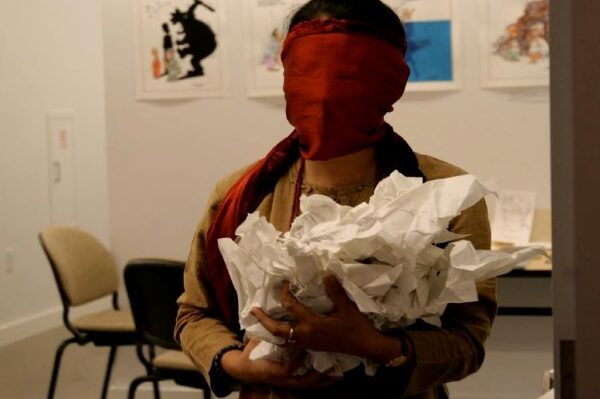 Artist masked standing in an office room space in a red scarf with an armful of crumpled white paper.