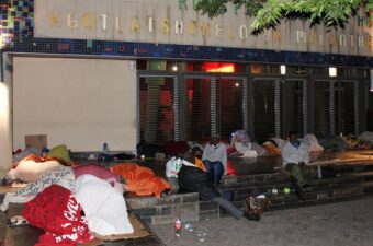 People resting and wrapped in blankets outside a building at night, some sitting and others lying down, with personal belongings scattered around.