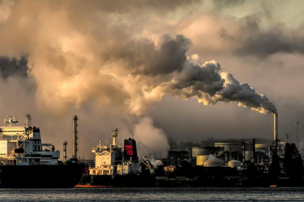 A large industrial facility emitting thick clouds of smoke into the sky, with ships docked nearby.