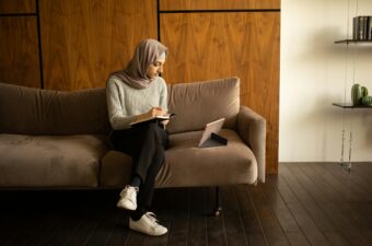 A muslim woman sits on a couch writing notes while watching her tablet.