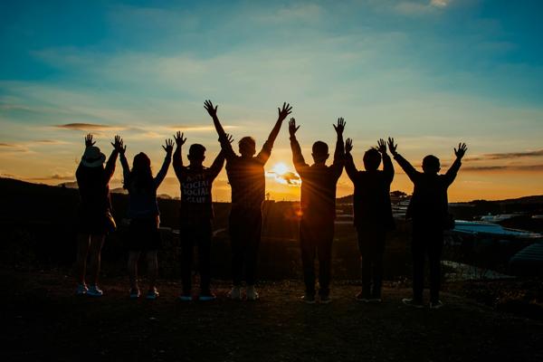 A group of diverse individuals standing on a hilltop with their arms raised towards the sky at sunset