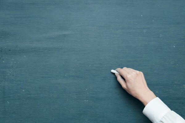 A hand holds a piece of chalk over a chalkboard.