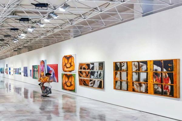 A person in a brightly lit art gallery observing a long wall filled with colorful abstract paintings. The spacious gallery has a polished floor reflecting the artwork and lighting, creating a modern and open atmosphere.