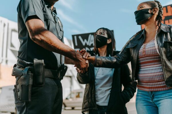 A Black woman and Asian woman are engaged in dialogue and holding the hand of a policeman with a sidearm.