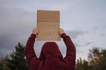 Person wearing a hooded sweatshirt holding up a cardboard sign that reads 'Love Shouldn't Hurt