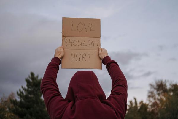 Person wearing a hooded sweatshirt holding up a cardboard sign that reads 'Love Shouldn't Hurt