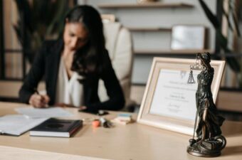 A woman sitting at a desk in a professional setting, focused on writing on a document. On the desk, there is a framed certificate, a book, and a statuette of Lady Justice holding scales