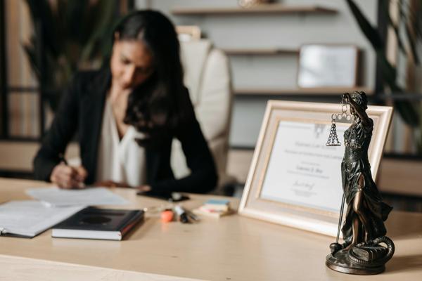 A woman sitting at a desk in a professional setting, focused on writing on a document. On the desk, there is a framed certificate, a book, and a statuette of Lady Justice holding scales