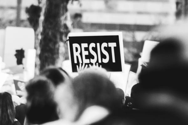 Black and white photo of a protest with a sign held up that reads 'RESIST' in bold letters, partially obscured by people in the crowd.