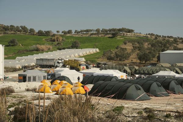 A humanitarian tent camp set against a green hilly landscape, featuring multiple rows of tents in various colors, trucks, and temporary structures