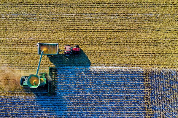 An aerial view of a tractor and harvesting machine working in a field, transferring crops into a container. The field shows patterns of harvested and unharvested sections,