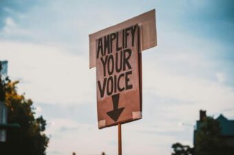 Handmade protest sign with the words 'Amplify Your Voice' written in bold letters, held up against a cloudy sky background