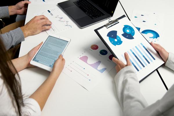 Group of people analyzing data charts and reports in a business meeting, with hands holding documents and digital devices.